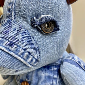 Puede incluir: Un peluche confeccionado con tela de mezclilla azul claro con costuras y texturas visibles. El ojo es un botón metálico con la palabra PLACE grabada. El juguete presenta orejas de tela marrón y un pequeño botón metálico color cobre en el pecho.