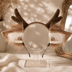 Puede incluir: Una diadema con astas de fieltro marrón y orejas de piel sintética suave en tonos beige, crema y blanco. Las orejas están situadas en la base de las astas y la diadema se muestra sobre un soporte circular de acrílico transparente.