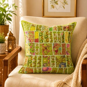 Puede incluir: Un cojín cuadrado con un diseño de patchwork compuesto por varios paneles de tela verde. La superficie está decorada con intrincados bordados, motivos florales y abalorios, unidos por costuras de color verde claro visibles en toda la pieza.