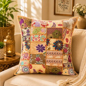 Puede incluir: Un cojín cuadrado con un diseño de patchwork compuesto por varios paneles de tela bordados. La superficie muestra una mezcla de patrones florales, de cachemira y geométricos en tonos vibrantes de morado, verde, azul, rosa y dorado sobre una base beige.