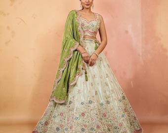 May include: A cream-coloured lehenga set featuring a sleeveless blouse and a voluminous skirt with intricate floral embroidery in pink, blue, and gold. An olive green dupatta with a scalloped, embroidered border is draped over one shoulder.
