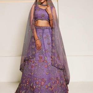 May include: A purple lehenga choli set featuring a sleeveless blouse and a floor-length skirt, both adorned with intricate floral embroidery, sequins, and beadwork. A sheer, matching purple dupatta with a thin border drapes over the head and shoulders.
