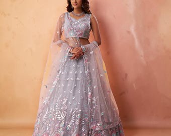 May include: A light grey lehenga choli set featuring a sleeveless blouse and a voluminous skirt made of net fabric. The ensemble is adorned with intricate silver embroidery, floral patterns in pink and teal, and a matching sheer dupatta with borders.