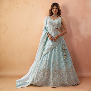 May include: A light blue lehenga choli set featuring intricate silver embroidery and beadwork. The sleeveless blouse has a sweetheart neckline, paired with a voluminous floor-length skirt and a matching dupatta draped over the shoulder.