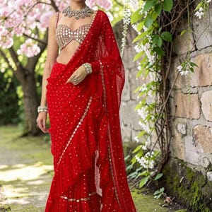 May include: A vibrant red saree featuring a ruffled hem and scattered gold embellishments. The ensemble includes a sleeveless, sweetheart-neckline blouse adorned with intricate gold mirror work, paired with a matching statement necklace and bracelets.