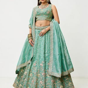May include: A seafoam green lehenga set featuring a sleeveless blouse and a floor-length skirt, both adorned with intricate floral embroidery in pink and gold thread. A matching sheer dupatta with a decorative border drapes over the shoulder.