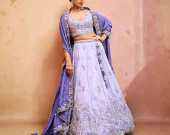 May include: A lavender lehenga set featuring a sleeveless crop top and a floor-length skirt, both adorned with intricate silver floral embroidery. A matching lavender dupatta with a wide, heavily embroidered silver border is draped over the shoulders.
