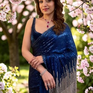 May include: A deep blue saree featuring dense sequin embellishments throughout the fabric, transitioning into a silver sequined border at the hem. The garment is paired with a matching sleeveless blouse and accessorized with a silver necklace.