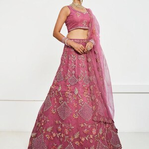 May include: A dark pink lehenga choli set featuring a sleeveless blouse and a floor-length skirt with intricate floral embroidery, beadwork, and sequin detailing. A matching sheer pink dupatta is draped over the shoulder and arm.