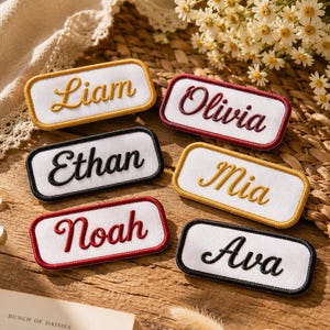 May include: Six rectangular white fabric patches with embroidered names in cursive script. The names Liam, Olivia, Ethan, Mia, Noah, and Ava are stitched in gold, dark red, and black thread, each framed by a matching colored border.