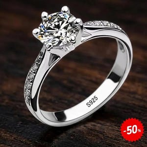 May include: A silver-toned ring featuring a large, round, faceted clear stone held by a six-prong setting. The band is accented with small, embedded clear stones along the sides and is clearly stamped with the text S925 on the inner surface.