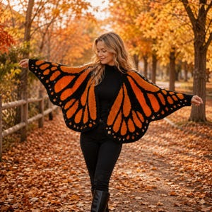 May include: A crochet shawl designed to resemble monarch butterfly wings, featuring a vibrant orange and black pattern with scalloped edges. The garment is worn over a black long-sleeved top and black pants, creating a striking contrast.