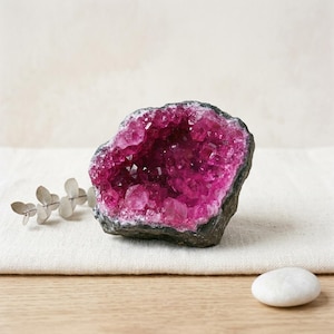 Könnte beinhalten: Eine leuchtend fuchsiafarbene Kristallgeode mit einer dunklen, rauen Außenhülle. Das Innere ist mit zahlreichen kleinen, funkelnden, durchscheinenden magentafarbenen Kristallen gefüllt. Ein kleiner Zweig getrockneter Eukalyptus und ein glatter weißer Stein liegen daneben.