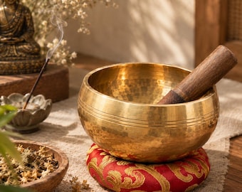 Peut inclure: Un bol chantant en métal doré avec une texture martelée et des lignes horizontales gravées, posé sur un coussin rond rouge à motif floral doré. Un maillet en bois est placé à l'intérieur du bol, posé sur une surface en tissu texturé.