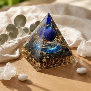 Peut inclure: Une pyramide en résine transparente contenant deux pierres sphériques bleu foncé, un fil métallique doré en spirale et une base de pierres noires mélangées à des flocons métalliques dorés. Un motif géométrique circulaire bleu est visible.