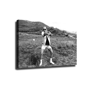 May include: A black and white photograph of a person wearing sunglasses, a light-colored jacket, and shorts, standing in a grassy field while holding a handgun with both hands extended forward. The background features rolling hills and sparse vegetation.