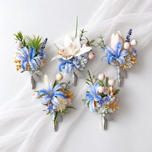 May include: Five floral boutonnieres featuring blue lilies, white orchids, pale pink tulips, small white berries, green foliage, and yellow accents. Each arrangement is wrapped with a silver ribbon stem, creating a cohesive set of decorative floral pins.