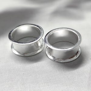 May include: A pair of silver-colored ear tunnels with a smooth, polished finish. These circular body jewelry pieces feature a hollow center and flared edges on both sides, designed for stretched ear piercings with a clean, metallic appearance.