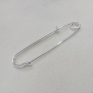 May include: A silver-toned metal safety pin featuring a classic elongated oval shape with a coiled spring mechanism at one end and a secure clasp closure at the other, designed for fastening fabric or securing various items together in a functional way.