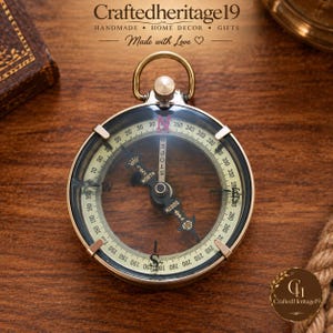Puede incluir: Una brújula redonda de latón con una cara de cristal transparente, una aguja central con marcas de Norte y Sur, y una escala de grados. La brújula tiene un anillo superior para colgar e incluye el texto Craftedheritage19 Handmade Home Decor Gifts.