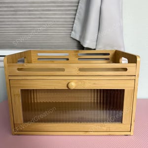 May include: A rectangular light brown wooden bread box featuring a top shelf with horizontal cutouts and a front-facing drop-down door. The door includes a central round wooden knob and a clear, vertically ribbed glass panel for visibility.