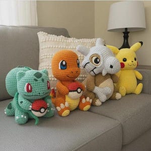 Op de afbeelding: Vier gehaakte knuffels bestaande uit een groene Bulbasaur met een rood-witte bal, een oranje Charmander met een rood-witte bal, een grijze Cubone met een schedelmasker en een gele Pikachu, op een rij geplaatst op een grijze bank.