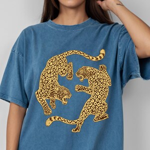 Könnte beinhalten: Ein blaues T-Shirt in Jeansoptik mit einem Grafikdruck von zwei goldenen Leoparden in kreisförmiger Anordnung. Die Leoparden sind in einer dynamischen Pose mit Flecken und geöffneten Mäulern dargestellt. Das T-Shirt hat einen Rundhalsausschnitt.