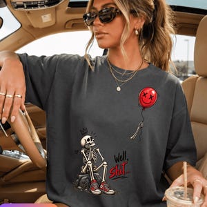 Peut inclure: Un t-shirt gris foncé à manches courtes avec un motif représentant un squelette assis sur un rocher, portant des baskets rouges et regardant un ballon rouge avec un visage souriant. Le texte "Well, Shit..." est imprimé en rouge et noir.