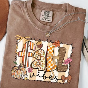 Op de afbeelding: Een bruin T-shirt met korte mouwen en een grafische print met de tekst "fall vibes". Het ontwerp bevat een pompoen, een eikel, herfstbladeren, strikken en ruitpatronen in de kleuren oranje, bruin en crème. Het shirt heeft een ronde hals.