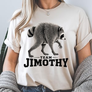 Pode incluir: Uma t-shirt de manga curta cor de creme com uma ilustração gráfica de um guaxinim cinzento e preto a caminhar para a direita. Abaixo do guaxinim, o texto "TEAM JIMOTHY" está impresso em letras maiúsculas pretas e a negrito no peito.