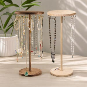May include: Two wooden jewelry stands with circular bases and tops. The left stand is dark brown wood holding multiple colorful beaded and metal necklaces. The right stand is light natural wood holding two necklaces, one with dark beads and one with white pearls.