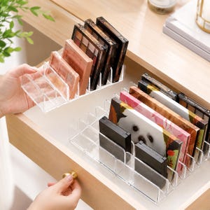May include: Two clear acrylic organizers with multiple vertical slots designed to hold rectangular makeup palettes upright. The organizers are positioned inside a wooden drawer, containing various palettes with different color schemes and designs.