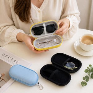 May include: A bright yellow oval zipped hard-shell glasses case containing black sunglasses and gold-rimmed spectacles. A light blue closed case and an open black case are nearby, each featuring a metal carabiner clip for secure attachment.
