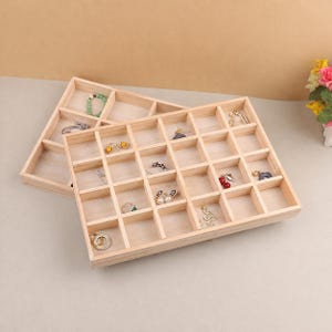 May include: Two light-colored wooden jewelry organizer trays with multiple square compartments. The trays are stacked at an angle and contain various pieces of jewelry, including rings, earrings, and a beaded bracelet, distributed across the individual grid sections.