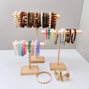May include: Three light-colored wooden T-bar jewelry stands displaying numerous beaded bracelets in various colors including white, blue, pink, green, yellow, brown, and black. A gold-tone starfish cuff and a white and gold bangle sit below.
