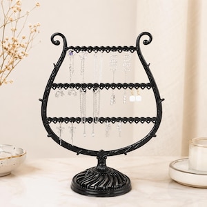 May include: A black jewelry stand shaped like a lyre with three horizontal bars featuring small holes for hanging earrings and necklaces. The stand has a circular, ribbed base and decorative scrollwork at the top, providing a structured display.