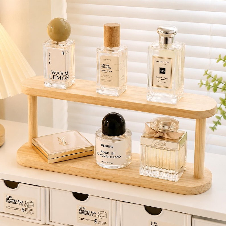 May include: A two-tiered light wood display shelf holding five glass perfume bottles and a square gold compact. The top shelf features bottles labeled Warm Lemon, Eau de Cologne, and Jo Malone. The bottom shelf holds a Beoupe bottle and a Chlo&eacute; bottle.