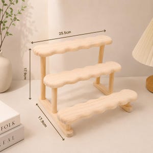 May include: A light-colored wooden three-tiered display stand with scalloped edges on each shelf. The stand features a width of 25.5 cm and a height and depth of 17.9 cm as indicated by the measurement lines printed on the image.