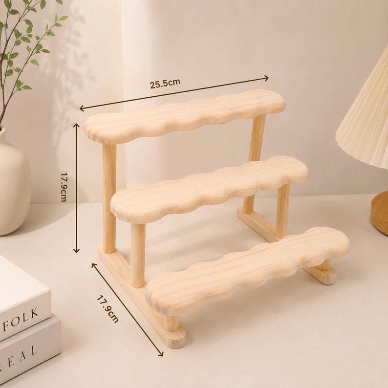 May include: A light-colored wooden three-tiered display stand with scalloped edges on each shelf. The stand features a width of 25.5 cm and a height and depth of 17.9 cm as indicated by the measurement lines printed on the image.