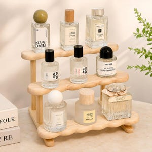 May include: A three-tiered wooden display stand with wavy edges, holding eight glass perfume bottles of various shapes and sizes. The bottles feature different caps, including round, cylindrical, and square designs, with labels displaying text.