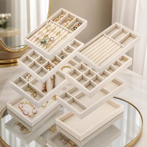 May include: A set of modular beige velvet jewelry organizer trays with various compartments, including ring rolls and square sections, containing assorted gold-toned rings, bracelets, and necklaces arranged in a stacked, tiered display configuration.