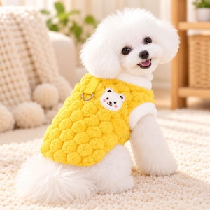 May include: A small white dog with fluffy fur wearing a bright yellow quilted vest. The vest features a textured, bubble-like pattern, a white fleece-lined collar, and a small embroidered bear patch on the chest with a metal D-ring attachment.