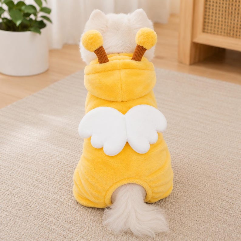 May include: A bright yellow plush pet hoodie featuring two brown-stemmed antennae with yellow pom-poms on top and a pair of white padded wings attached to the back. The soft, fuzzy garment is designed to cover the torso and head of a small animal.