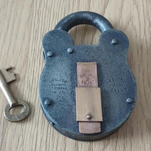 May include: A dark grey metal padlock with a rounded shackle and a brass-coloured central plate featuring the text SECURE 2 LEVER. The lock body is embossed with ENGLISH PATENT N 525346 and REGD BRAND. A matching brass-coloured key lies to the left.