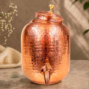 May include: A cylindrical copper water dispenser featuring a hammered texture, a matching lid with a small knob handle, and a metal spigot attached to the lower front section. The vessel has a polished metallic finish and a rounded body shape.