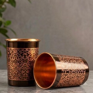 May include: Two cylindrical tumblers made of polished copper with a dark brown finish. Each tumbler features an intricate, embossed floral scrollwork pattern that reveals the bright copper colour beneath the dark surface.