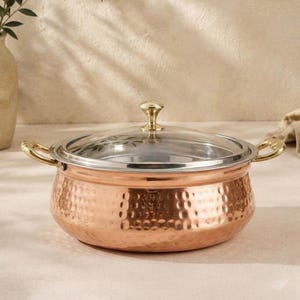 May include: A round, hammered copper cooking pot with a polished metallic finish. It features two curved side handles and a clear glass lid topped with a small, rounded gold-toned knob. The pot has a wide, shallow base and a smooth interior rim.