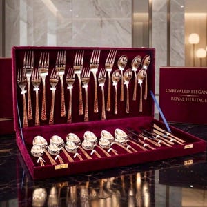 May include: A flatware set in a deep red velvet-lined case with the text UNRIVALED ELEGANCE ROYAL HERITAGE. The set includes rose gold-toned forks, spoons, and knives with textured handles, arranged in rows within the open rectangular box.