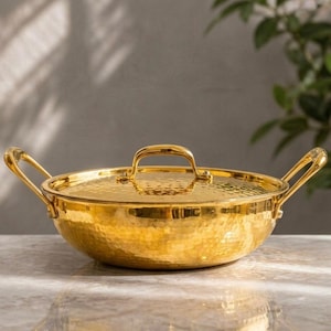 May include: A round, gold-colored metal serving dish with a hammered texture, featuring two curved side handles and a matching lid with a central arched handle. The polished surface reflects light, showcasing the dimpled, metallic finish.