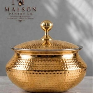May include: A gold-toned, hammered metal serving bowl with a matching lid and a decorative, ribbed spherical knob handle. The bowl features a wide, rounded body and a smooth, metallic finish. The text "MAISON PANTRY CO." is visible above.